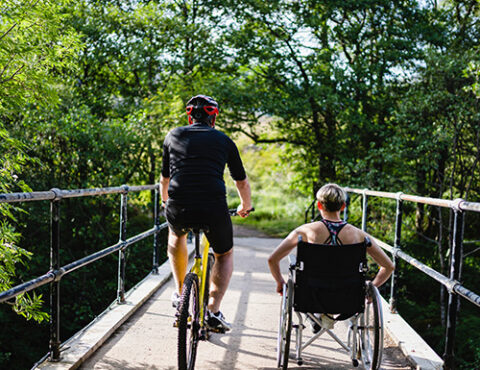 Man on a bike rides to the left looking at a lady in a wheelchair on the right passing across a bridge in a forest on a sunny day